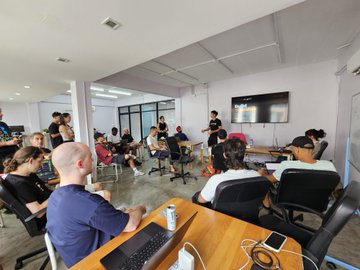A casual gathering of people sitting in a circle of chairs within a brightly lit room, with some individuals using laptops and engaging in conversation.