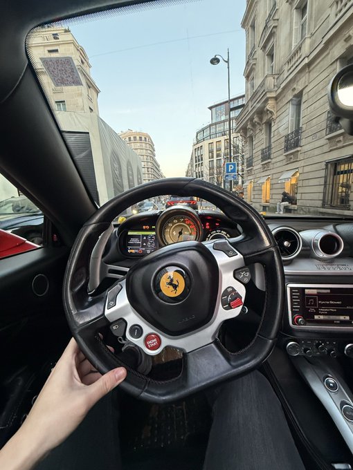 A first-person view from the driver's seat of a Ferrari shows a hand on the steering wheel with the prancing horse logo, looking out onto a European city street with classic architecture.