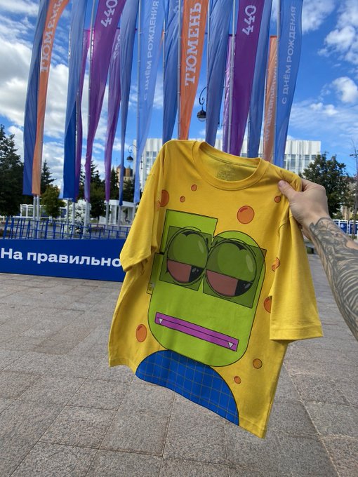 A hand holds up a yellow t-shirt featuring a green cartoon character with sunglasses and a pink mouth, set against an outdoor background with colorful Russian flags.