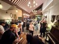 A crowded, dimly lit bar with patrons mingling under warm pendant lighting, capturing the lively atmosphere of a "Mahjong Night LA" event hosted by Bored Ape Yacht Club, where the energy matches the "vibes on ten thousand" sentiment.