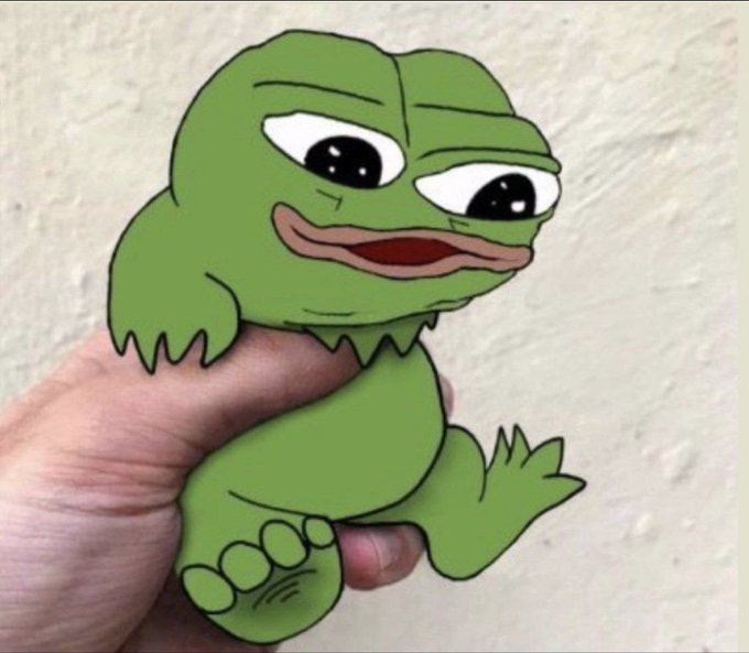 A hand holding a small paper craft of the Pepe the Frog meme character, showing his green face with large expressive eyes.