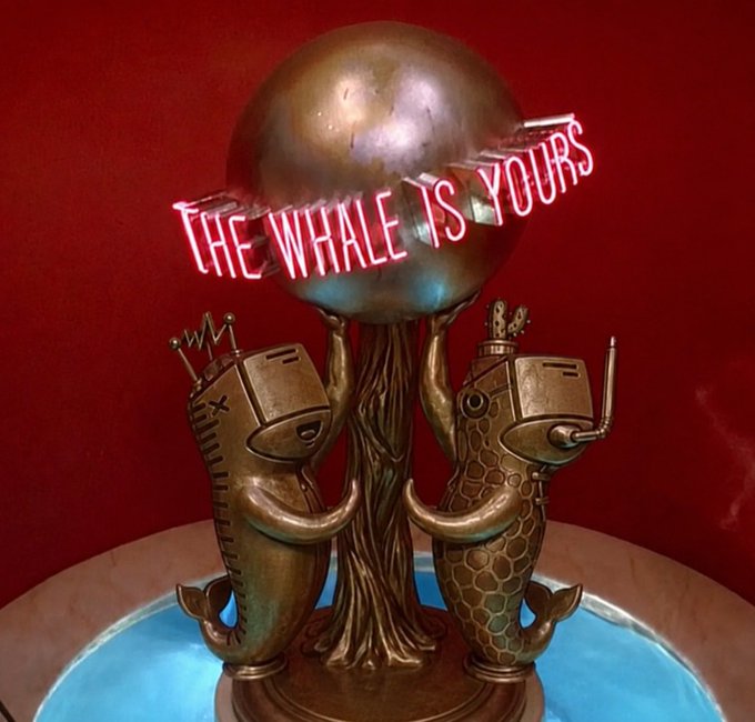 A bronze sculpture of a whale and a robotic figure holding up a sphere, with a neon sign reading 'THE WHALE IS YOURS' against a red wall.