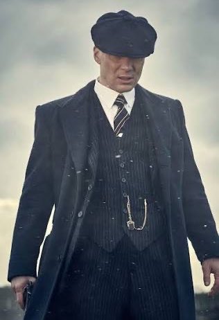 A stern-looking man in a dark pinstripe suit and flat cap stands against a cloudy sky, evoking the gritty aesthetic of a *Peaky Blinders* character. The image's somber, stormy mood mirrors the post's coverage of the crypto market's recent downturn, with Bitcoin slipping below $63k amid broader macroeconomic concerns.