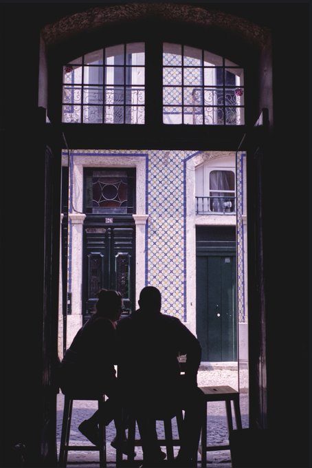 A silhouette of two figures on stools framed by a dark arched doorway, looking out toward a sunlit Lisbon street with blue and white tiled buildings.