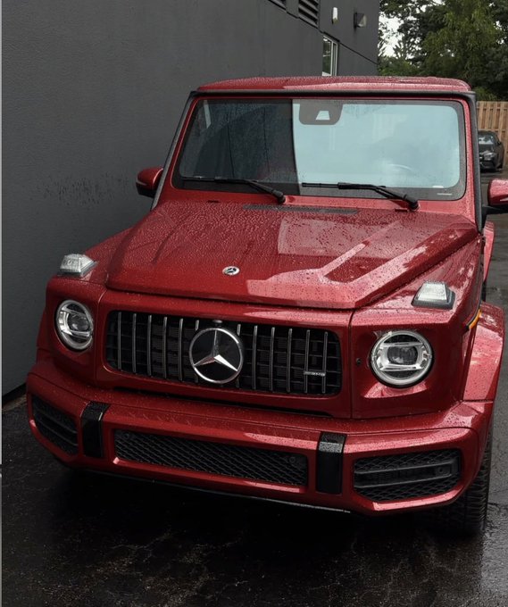 A red Mercedes-Benz G-Wagon parked in front of a dark building, its glossy finish beaded with raindrops.