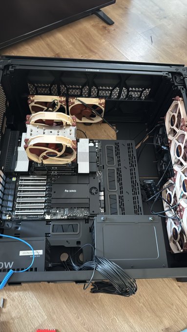 A custom PC build in progress sits on a wooden floor, featuring a Pro Series motherboard, Noctua fans with copper heatsinks, and visible "PRO SERIES" branding, with loose cables still being organized—reflecting the "slowly but surely" progress of the assembly.