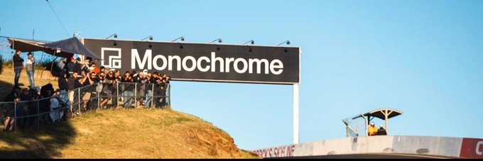 A crowd of spectators leans against a railing on a hill, watching an event beneath a large black sign that reads 'Monochrome' with a stylized 'M' logo.