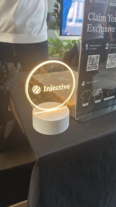 An illuminated circular 'Injective' branded neon sign on a white stand sits on a black table next to a QR code display and merchandise.