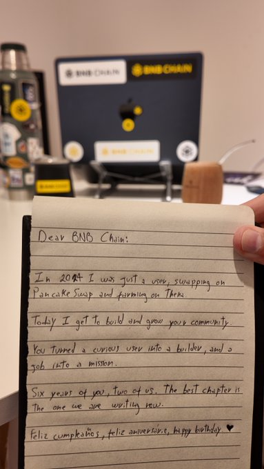 A handwritten note on lined paper is held in front of BNB Chain merchandise, with the message 'Dear BNB Chain: In 2019 I was just a noob swapping on PancakeSwap and farming on Thorus. Today I get to build and grow your community... Feliz cumpleaños, feliz aniversário, happy birthday.'
