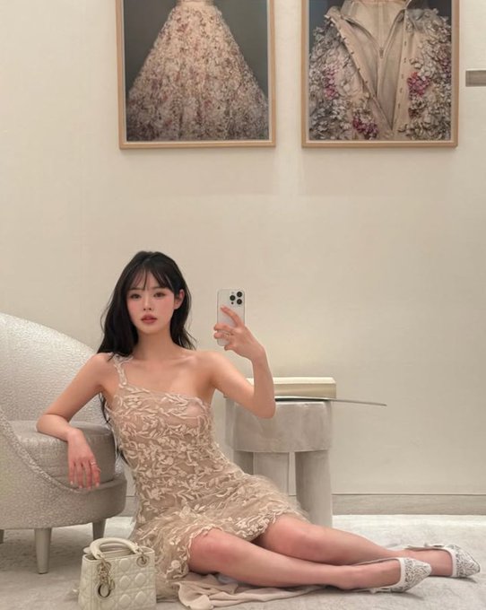 A young woman takes a mirror selfie while sitting on the floor in a beige floral dress, holding a phone, with a small white handbag beside her and two framed floral artworks hanging on the wall behind her.