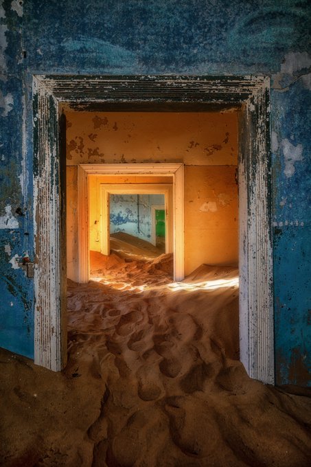 A photograph of an abandoned building interior with peeling blue paint, showing a doorway leading through rooms that have been filled with sand dunes, with warm light illuminating the space.