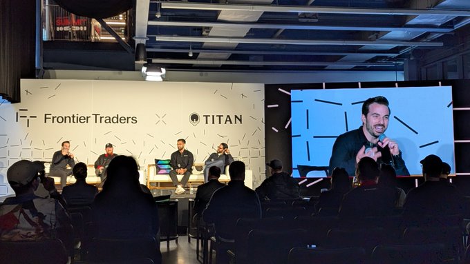 A panel discussion on stage at an event branded 'Frontier Traders' and 'TITAN,' with four speakers seated in front of an audience and one speaker shown on a large screen.