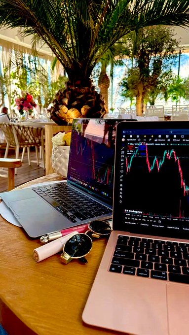 A laptop and tablet on a wooden table display cryptocurrency trading charts, with sunglasses, a lipstick, and a tropical plant in the background.