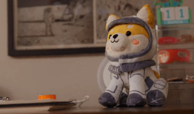 A plush Shiba Inu toy dressed in a gray and white astronaut suit sits on a table near a calendar showing November 11.