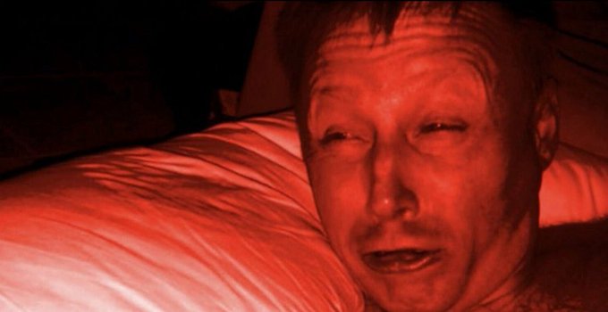 A close-up of a man's face bathed in a deep red hue, resting against white pillows in a dimly lit setting.