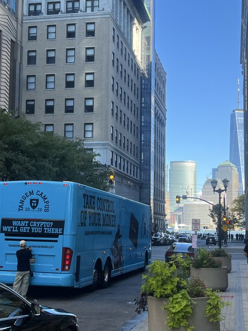 A blue bus advertising 'TANGEM CAMPUS', 'WANT CRYPTO? WE'LL GET YOU THERE', and 'TAKE CONTROL OF YOUR MONEY' is parked on a city street, with One World Trade Center visible in the background.