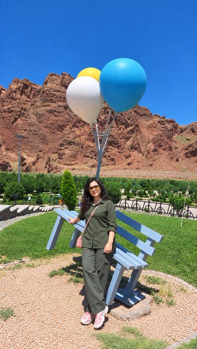 A woman in dark green attire and sunglasses stands on a gravel path next to a blue bench, holding a bunch of yellow, white, and blue balloons with a rocky red mountain landscape behind her.