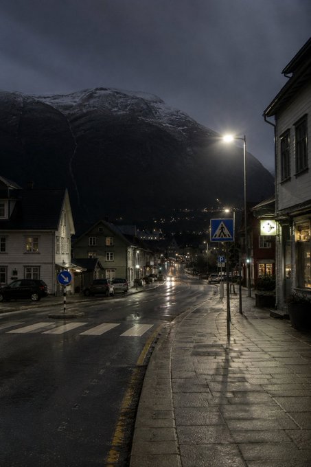 A nighttime photograph of a wet street in what appears to be a Scandinavian or northern town, with a snow-capped mountain looming in the background, illuminated street lamps, and a pedestrian crossing sign.