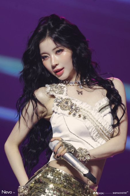 A female performer with long black hair performs on stage wearing a white embellished crop top and gold sequined skirt, holding a microphone.