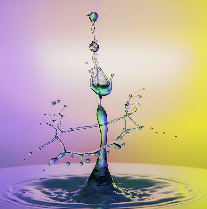 A vibrant water droplet collision captured mid-splash against a gradient background transitioning from soft purple to bright yellow.