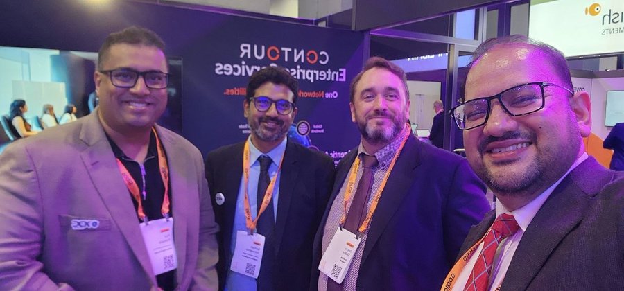 Four men in business attire posing for a selfie at what appears to be a technology event, with signage reading 'COCKTAIL' and 'Enterprise' services visible in the background.