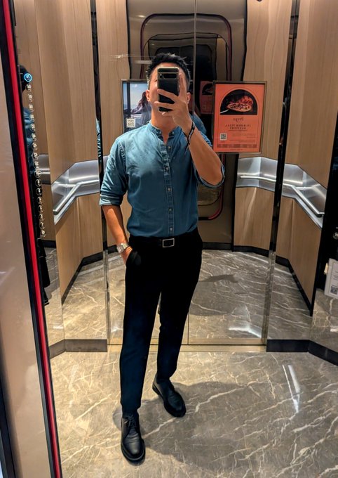 A man in a teal button-up shirt, dark trousers, and black dress shoes takes a mirror selfie inside a polished elevator with marble flooring, posing with one hand in his pocket. Behind him, a framed poster advertises a "CHEERS TO ALL LUCKY STARS" event, tying into the post about his evening at the Coinbase advanced F1 qualifying watch party at SKAI Bar in Swissôtel The Stamford.