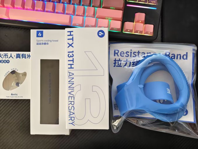 A top-down photo of sports accessories on a desk, including a pink keyboard, a white 'HTX 13TH ANNIVERSARY' sports cooling towel box, a crypto trading desk pad, and a blue resistance band in its packaging.