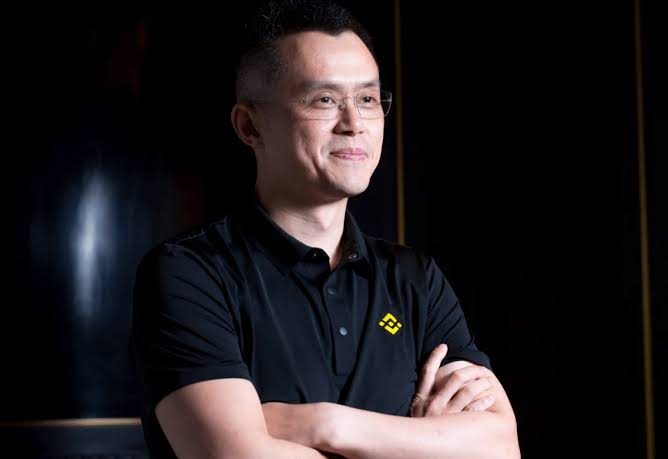 In the image, there's an Asian man wearing glasses and a black polo shirt with his arms crossed over his chest. He has a big smile on his face as he looks off into the distance. The background of the photo is dark, which makes him stand out even more. It appears that he might be posing for a picture or standing in front of an art piece related to Binance's tokenization system and America fighting it.