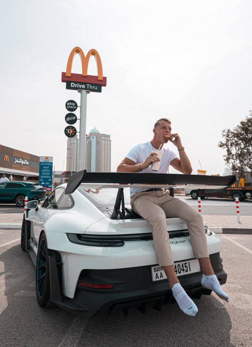 A young man casually sits on the rear spoiler of a white Porsche GT3 RS (Dubai plate "A 40451") eating McDonald's fries in front of a McDonald's Drive Thru sign, humorously illustrating the meme's punchline of Bitcoin investments transforming humble fast-food employment into luxury wealth.