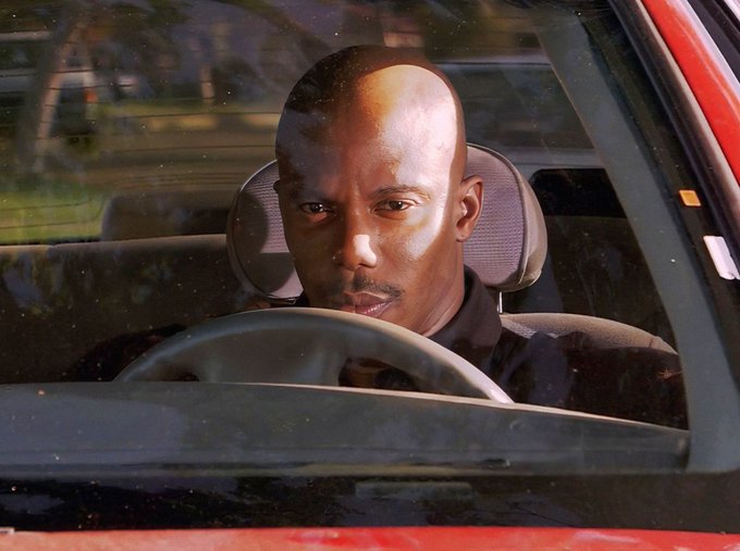 A bald Black man sits in the driver's seat of a red car, looking out the window with a serious expression.
