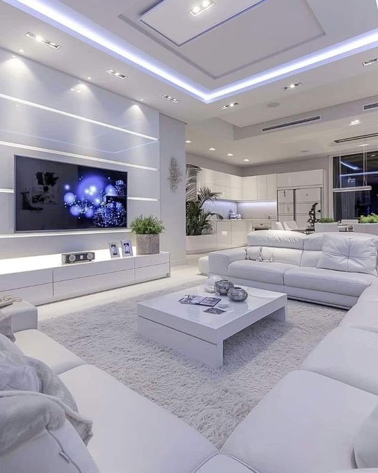 A modern, brightly lit white living room featuring a large wall-mounted TV displaying a blue abstract image, white leather sectional sofas, a white coffee table, and an open view into a contemporary kitchen.