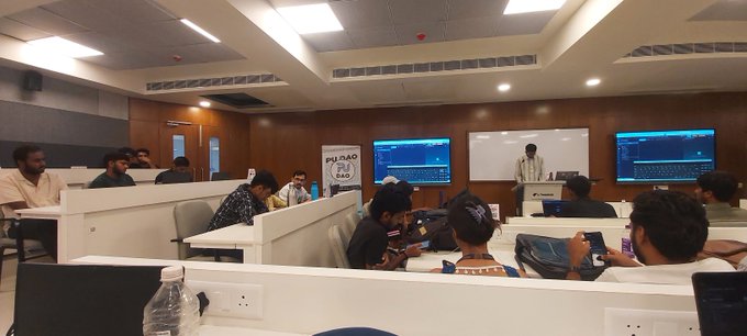 A classroom or training room scene with students seated at desks with monitors, while an instructor stands at the front near a whiteboard and screens, with a 'PUDU RAO RAO' banner visible.