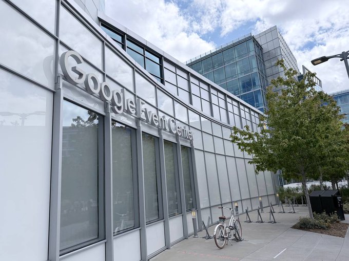 An exterior view of a modern glass building displaying the text "Google Event Center" on its facade, with a bicycle parked along the sidewalk.