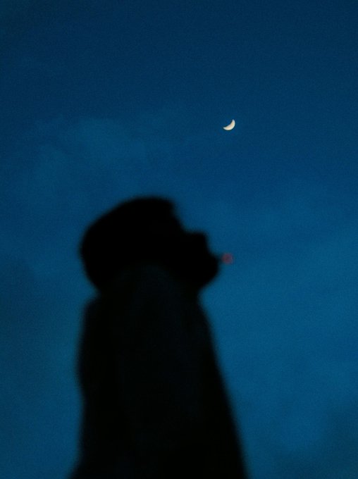 A silhouette of a person blowing smoke against a dark blue night sky, with a crescent moon visible in the background.