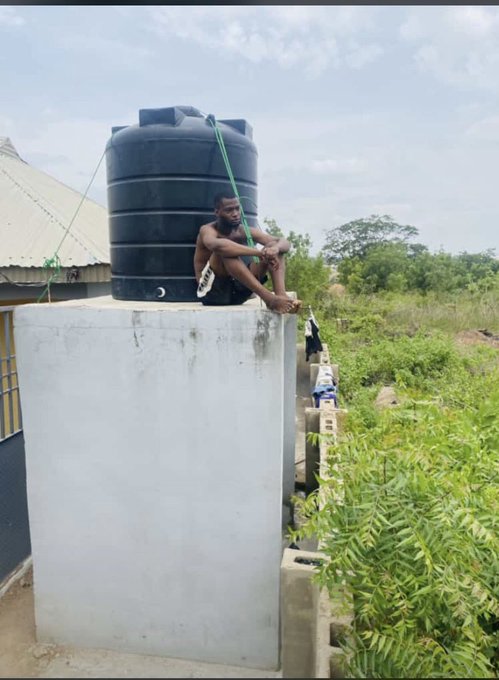 A shirtless man sits on a concrete water tank platform beneath a large black plastic water storage tank, with green vegetation and a metal fence visible in the background.