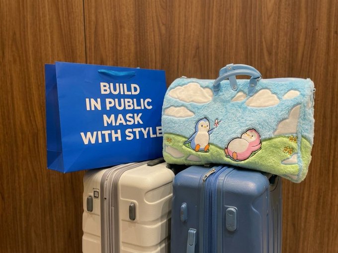 Two suitcases (one beige, one blue) topped with a blue fuzzy bag featuring cartoon penguins and clouds, alongside a blue shopping bag reading "BUILD IN PUBLIC MASK WITH STYLE".