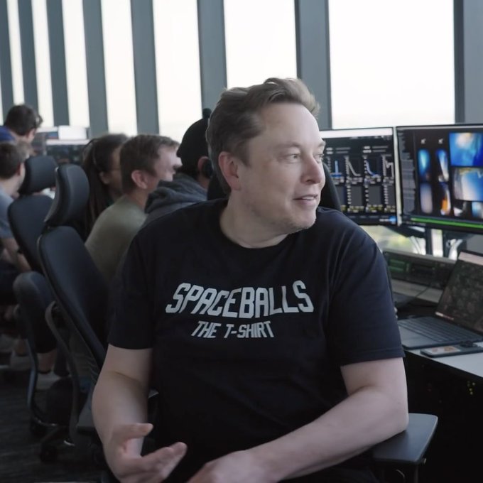 A man resembling Elon Musk wearing a black 'SPACEBALLS THE T-SHIRT' sits in an office environment with multiple computer monitors in the background.
