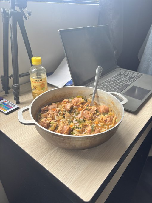 A pot of chicken and rice stir-fry sits on a desk next to a laptop and a bottle of "hafri" juice. The image illustrates the post's caption about finally making a long-awaited meal, inviting viewers to guess the ingredients for a fun challenge.