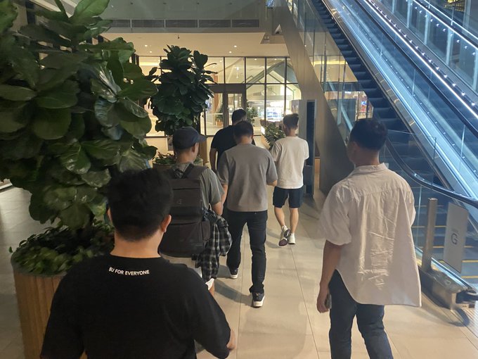 Several people walk away from the camera through a modern mall or hotel corridor, passing large potted fiddle-leaf fig plants near an escalator.