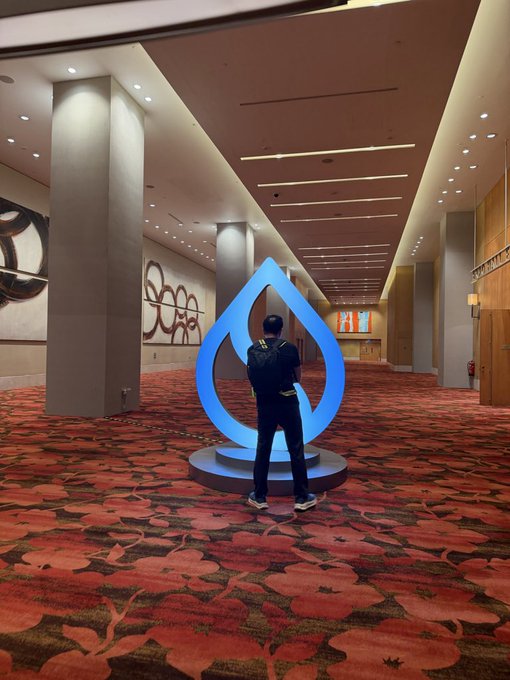A person with their hands behind their back stands in a large convention hall hallway facing a large blue droplet-shaped sculpture.