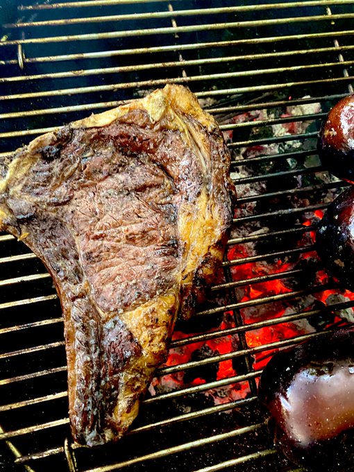 A close-up image of a large T-bone or ribeye steak cooking on a grill over glowing red-hot coals, with what appears to be sausages on the right side.