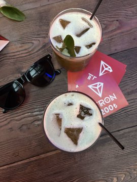 Two iced beverages garnished with mint leaves sit on a dark wooden table alongside black sunglasses and a pink card displaying the TRON logo and "27." text, connecting the post's mention of the TRON TGF VIP event held during the World Cup.