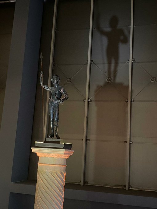 The image features a statue of a man holding a spear, standing tall on a pedestal or column. The statue casts a shadow onto the wall behind it, creating an interesting visual effect. The caption above the image reads "super hyped waking up to @TheARCTERMINAL updates this morning. the way ANIMA maintains the entire knowledge web across sessions is actually changing how i work in web3. no more losing flow when life interrupts." This suggests that the statue and its shadow are symbolic representations of the importance of maintaining a consistent online presence, even during challenging times or interruptions in our daily lives.