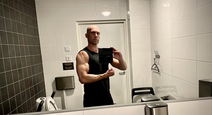 A muscular bald man wearing a black tank top takes a mirror selfie in a tiled bathroom while flexing his bicep.
