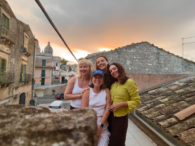 A group photo of four smiling women posing together on a rooftop at sunset, with an old Italian town and dome visible in the background.