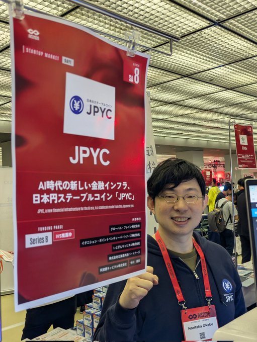 A smiling representative wearing a JPYC lanyard and nametag (Noritaka Okabe) poses in front of a red exhibition banner at Startup Market SA8 on the 1st floor of IVS, promoting the Japanese yen stablecoin JPYC (Series B, IVS exclusive) as a new financial infrastructure for the AI era.