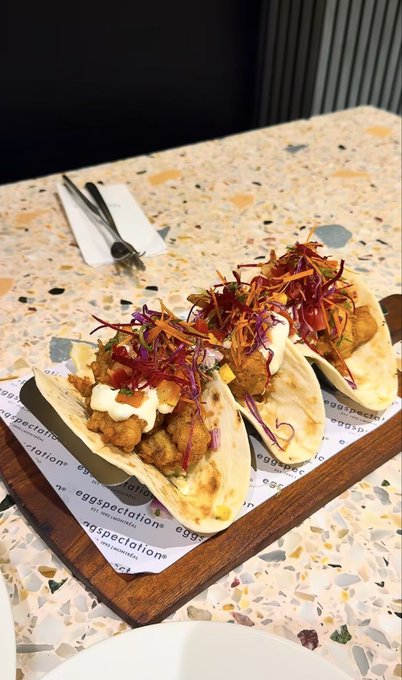 Three gourmet fried chicken tacos garnished with purple cabbage, red sauce, and microgreens are served on a branded "Eggspectation" paper-lined wooden board, resting on a terrazzo table. The post playfully asks viewers to guess the $SOL (Solana cryptocurrency) price of this upscale meal from the Eggspectation restaurant chain.