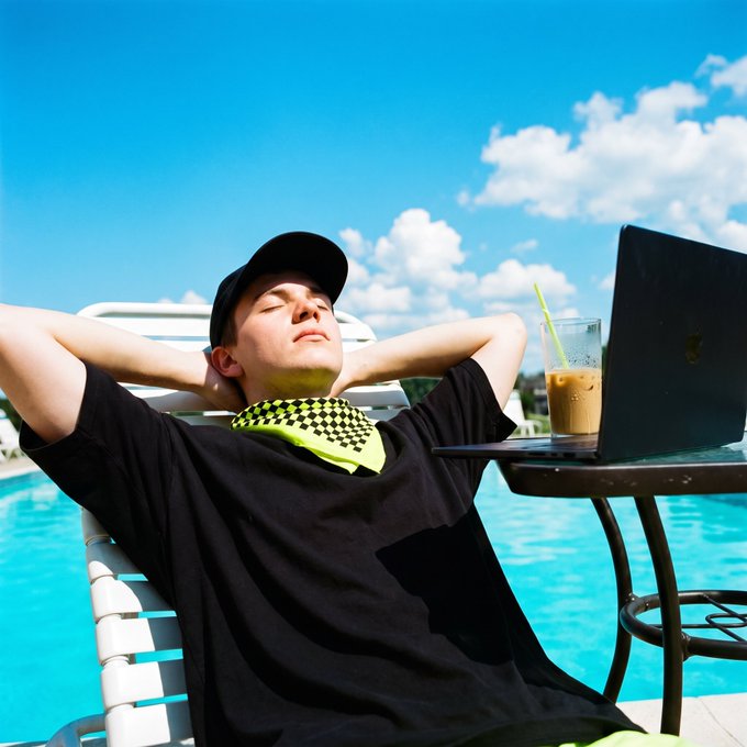 A young man relaxes in a chair by a swimming pool on a sunny day, wearing a black cap, black t-shirt, and a neon green bandana, with a laptop and iced coffee on a table beside him.