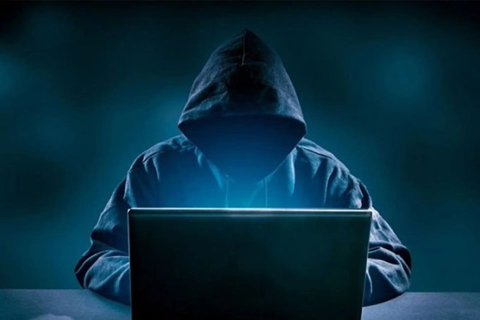 A mysterious hooded figure sits in front of a glowing laptop in a dark, blue-lit environment, face obscured by the hood.