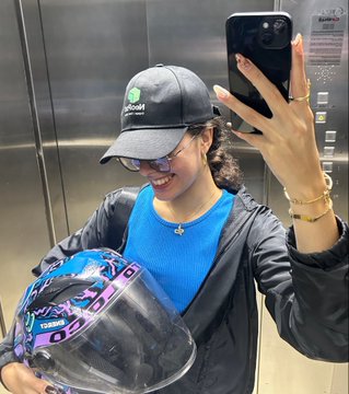A woman wearing a black cap, sunglasses, and a blue top takes a mirror selfie in an elevator while holding a blue, black, and purple motorcycle helmet.